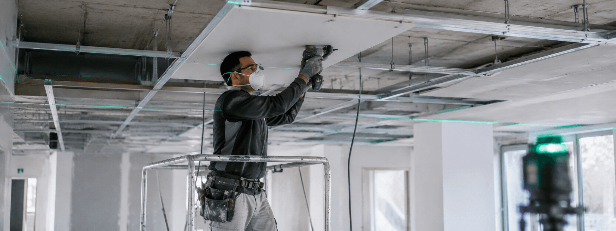 Plaquiste détaché européen en plein montage d'un faux plafond suspendu sur ossature métallique et suspentes, réglage de la planéité au niveau laser, plateau tertiaire, geste technique de précision